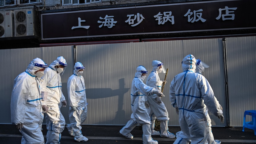 A group of men and women in white hazmat suits walk in front of Chinese lettered storefront in evening
