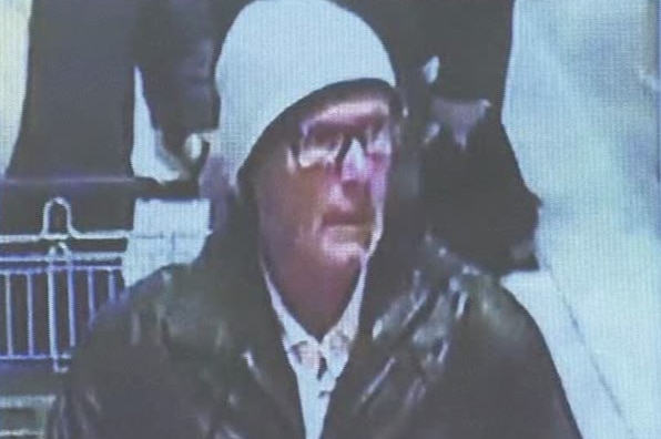 A man wearing a beanie and glasses is walking in front of a shopping trolley, looking away from the camera.