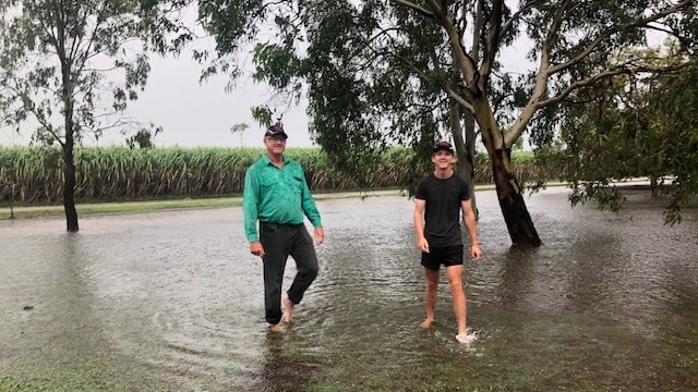 Two men wade through a flooded yard with their cane farm in the background