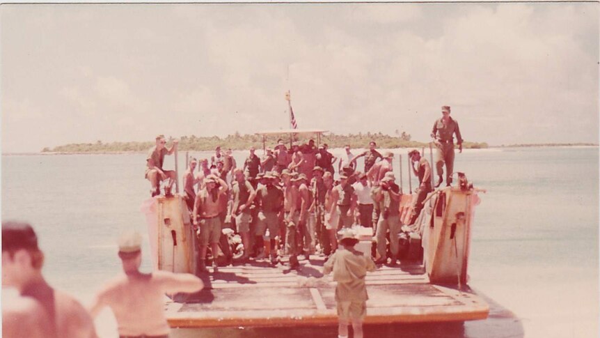 A group of soldiers, many shirtless stand on a boat as it makes landfall.