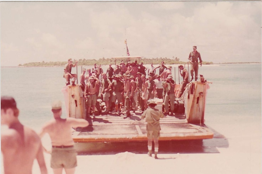 A group of soldiers, many shirtless stand on a boat as it makes landfall.