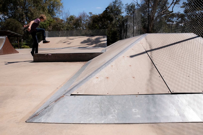 A wooden skate ramp is warping away from its fastenings as a teenager skates in the background.