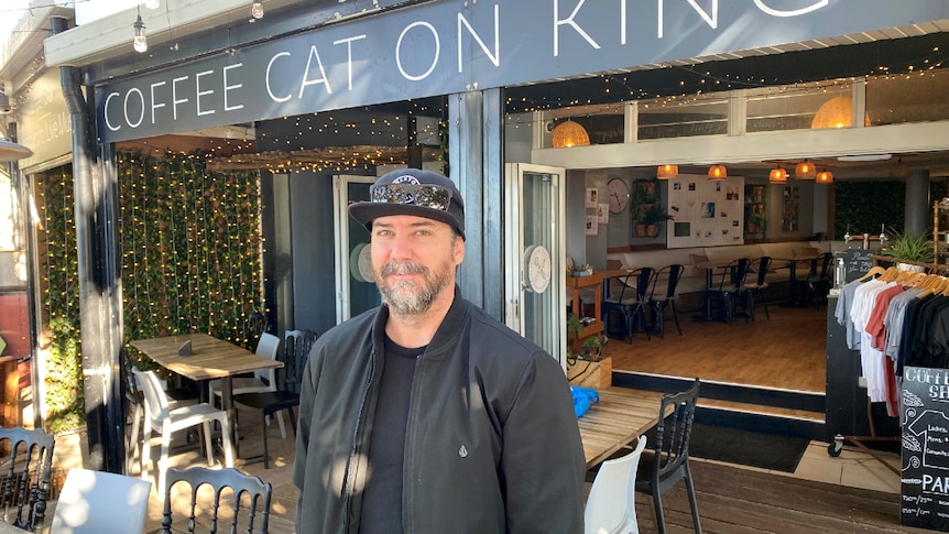 A man with a serious face, wearing a dark jacket, T-shirt and cap stands in front of the Coffee on Kings shop