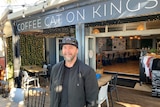 A man with a serious face, wearing a dark jacket, T-shirt and cap stands in front of the Coffee on Kings shop