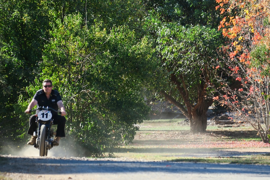 Peter rides without a helmet up a dirt track, small trees line the road.