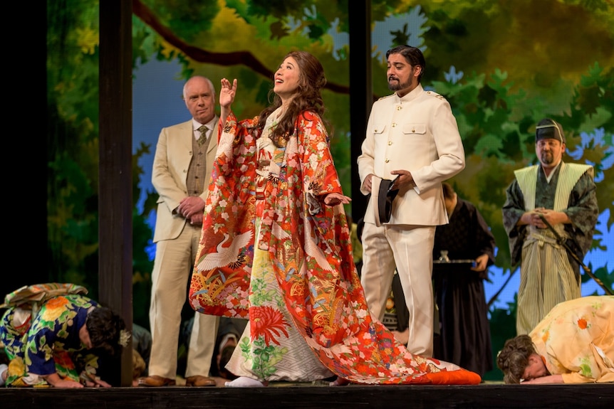 A photo of an opera where a woman in colourful Japanese dress sings, with several men looking on