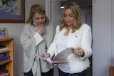 Two women, one in a grey jumper and the other in a white suit, stand in a consulting room looking at a document