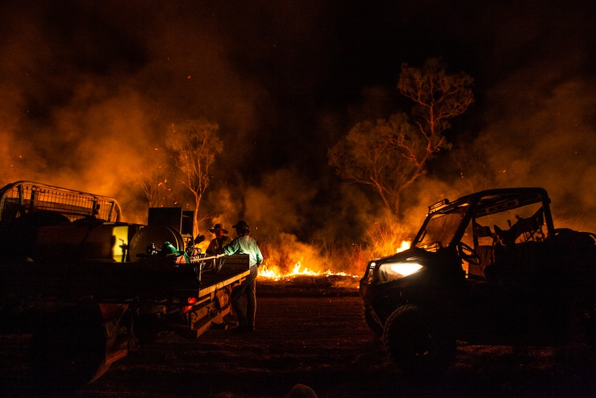 Two water trucks parked in front of a line of flames in bushland at night.