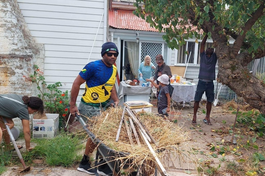 Man carries a full wheelbarrow. Woman weeds to his right. Behind him people are working hard in yard. 