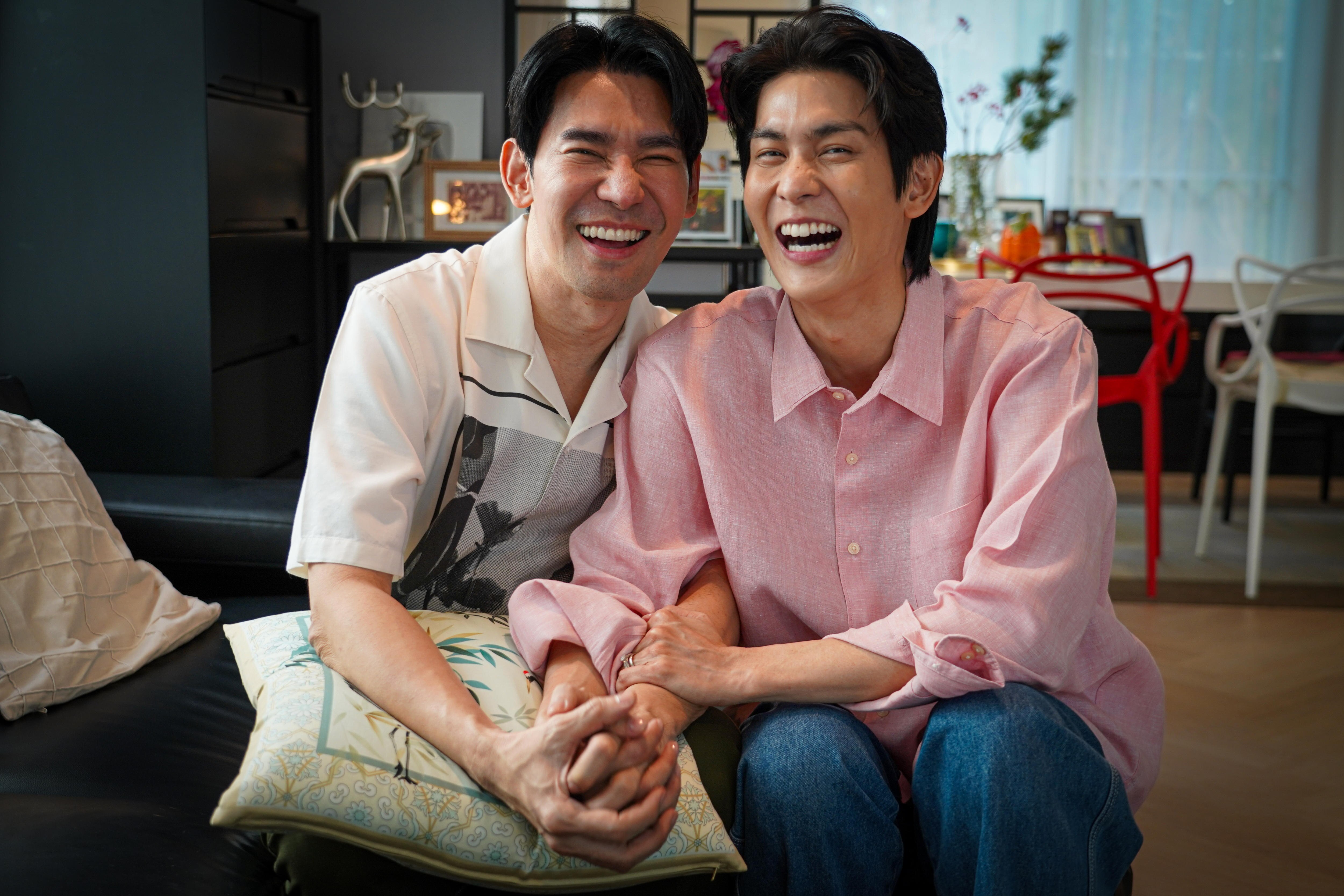 Two men with black hair smile widely while holding each others arms and hands while looking at the camera