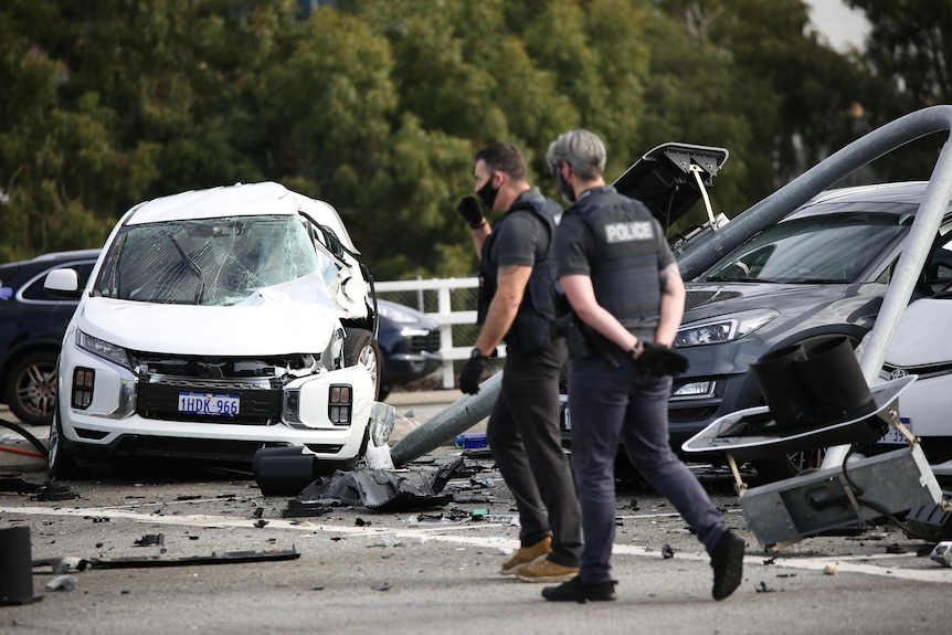 Two police officers in plain clothes and police vests walk past the scene of a crash with a pole down over the road.