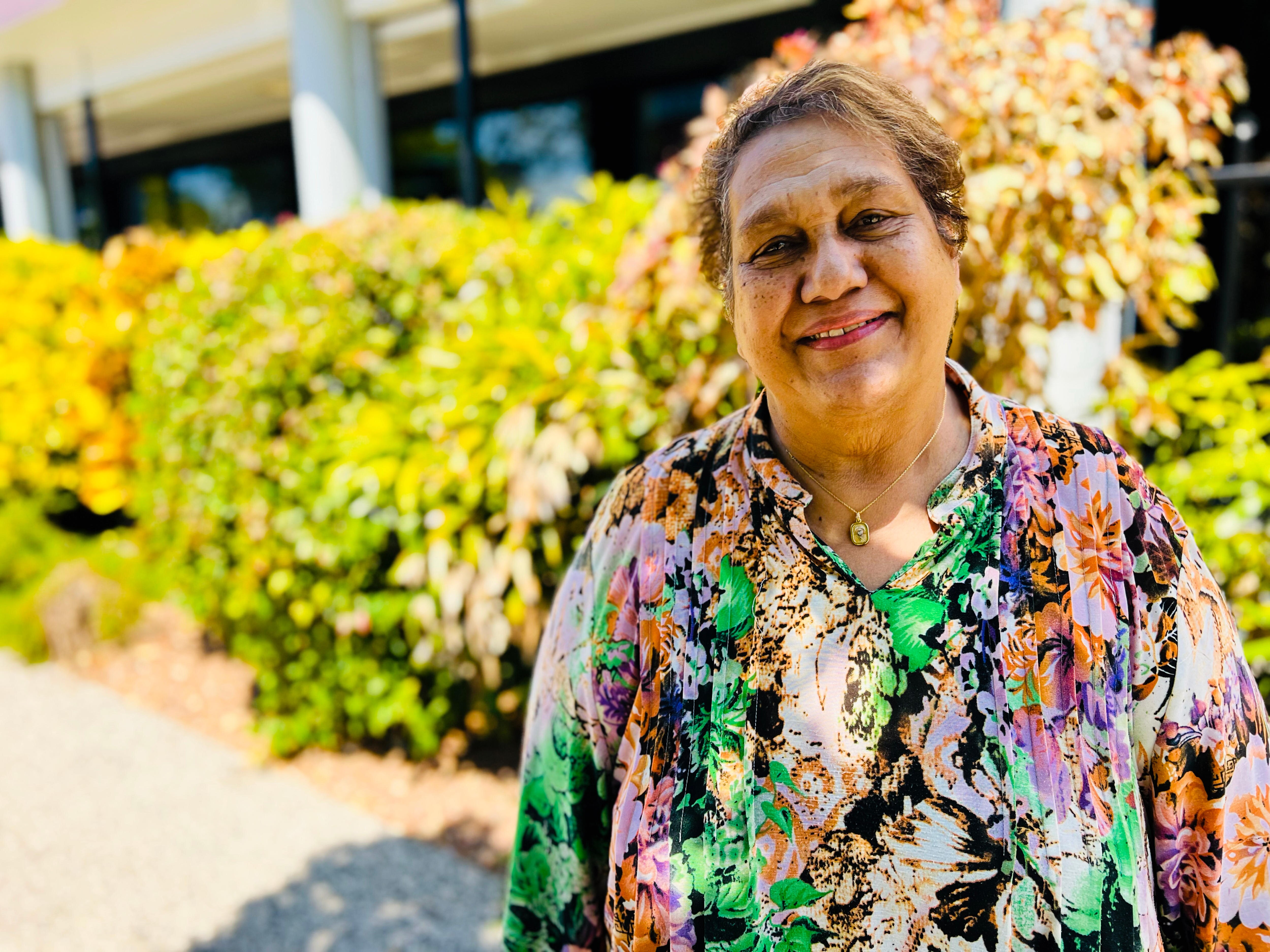 A lady stands on a side walk in front of a row of large plants as she smiles at the camera face on