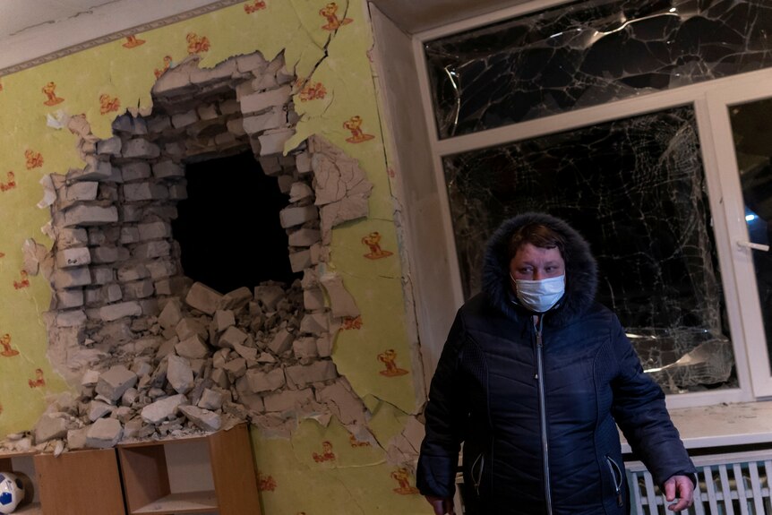 A person in a face mask and hooded jacket stands next to a hole blown in a wall in Ukraine.