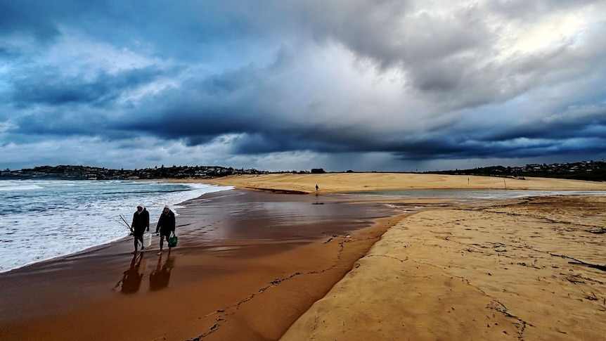 Two people walking on the beach in Sydney with rainclouds looming above.