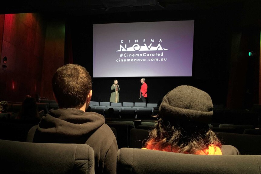 The backs of a man and a woman's head looking at a cinema screen with two people on stage.