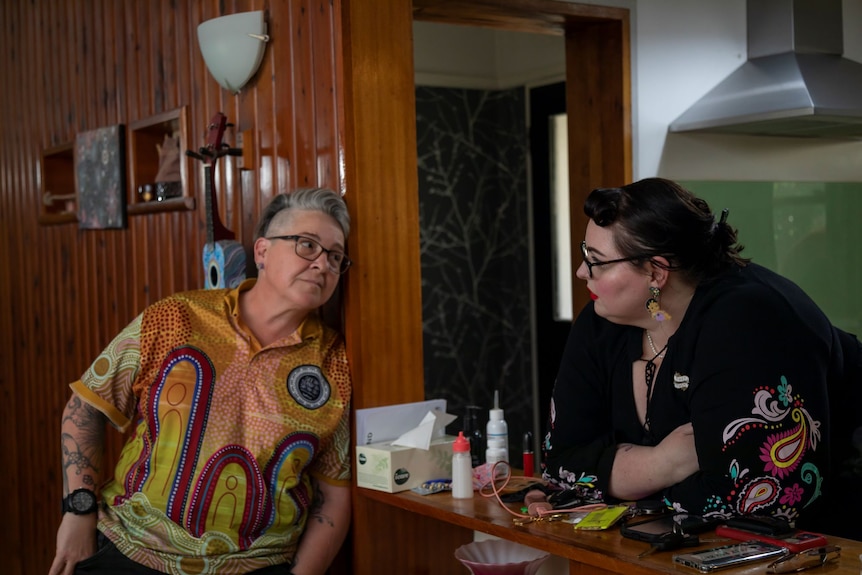 A woman with short grey hair leans against a wall wearing an Indigenous Tshirt, looking at her wife who wears black.