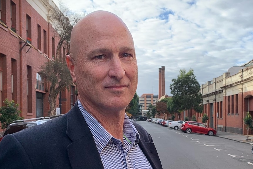 A bald man wearing a suit jacket and shirt stands in a quiet street.