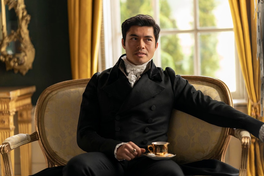 A man in formal clothing sits in a broad armchair, balancing a cup of tea on his lap.