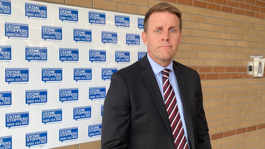 A man in a suit stands against a wall with crime stoppers logos.