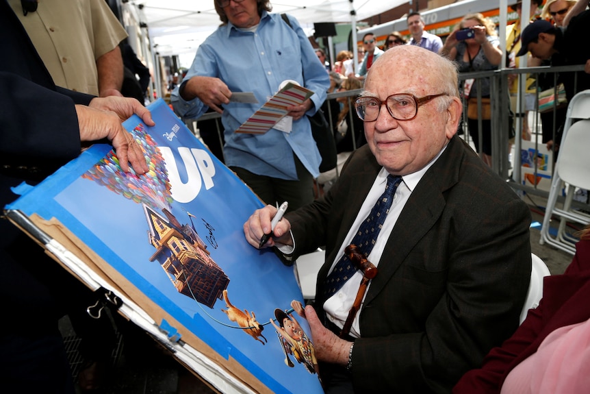 Ed Asner signs a poster while sitting down