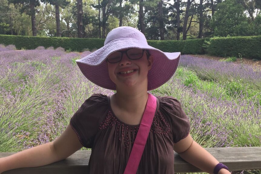 A girl wearing a purple floppy hat and black glasses stands in a lavender field and smiles.