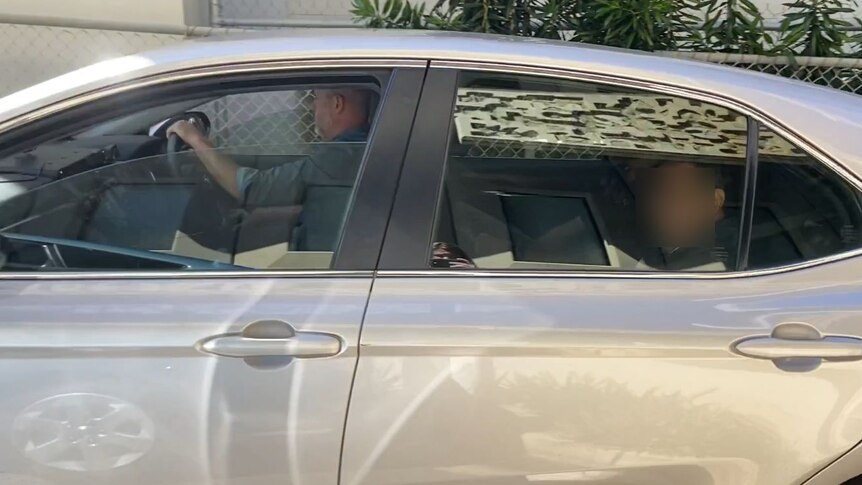 Man in back seat of car is driven to court with face blurred.