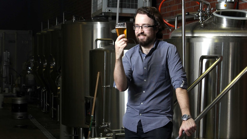 Craft beer brewer Phil O'Shea holds a glass of beer, standing in front of beer production equipment.