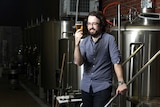 Craft beer brewer Phil O'Shea holds a glass of beer, standing in front of beer production equipment.