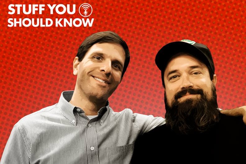 The two hosts of the Stuff You Should Know podcast smile in a promotional photo against a bright red background.