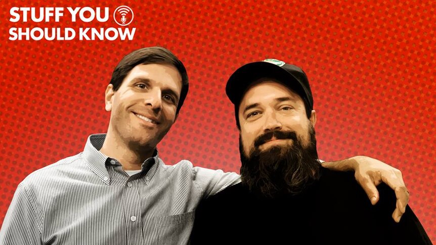 The two hosts of the Stuff You Should Know podcast smile in a promotional photo against a bright red background.