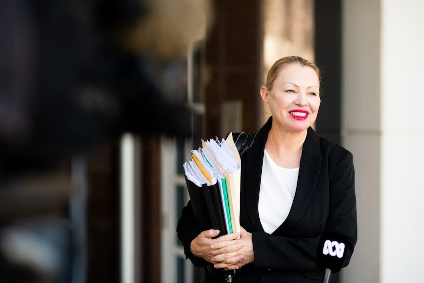 A woman in a black suit and white shirt stands in front of a microphone. She holds paperwork.