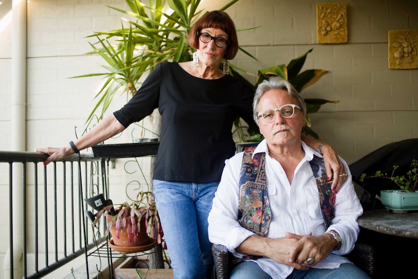 A woman with short hair wearing thick-rimmed glasses stands with a serious expression next to a man seated, wearing glasses