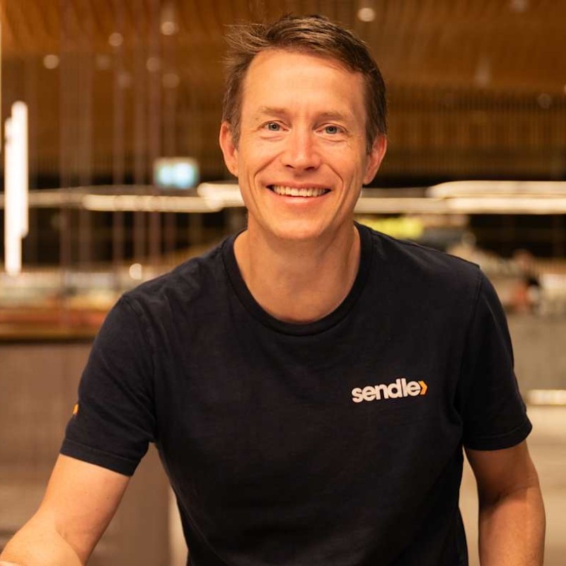 El sonriente James viste una camiseta negra con la etiqueta Sendler y el fondo está borroso.