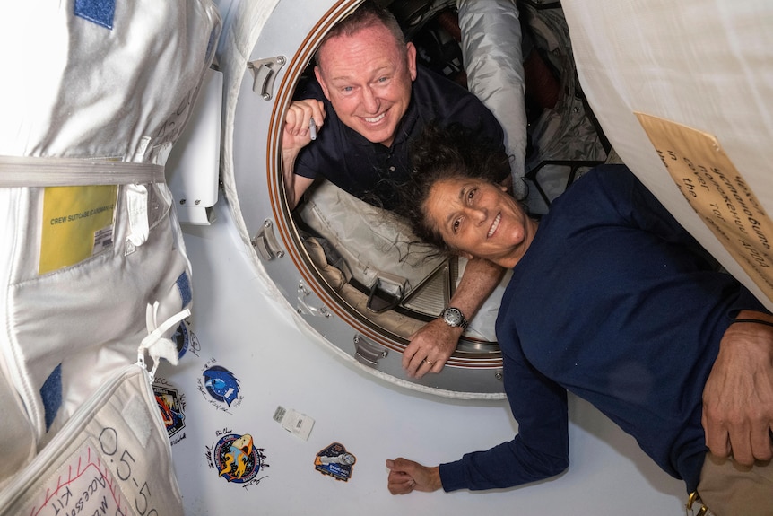 A middle aged white man and a woman with dark curly hair pose by a round port door inside a space station