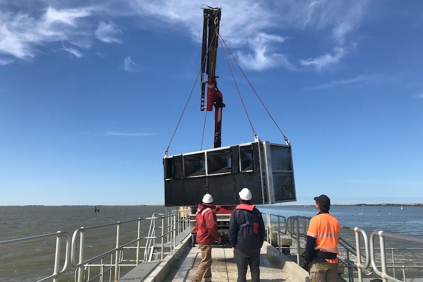 Three man and a crane lifting up a fish cage from the ocean.