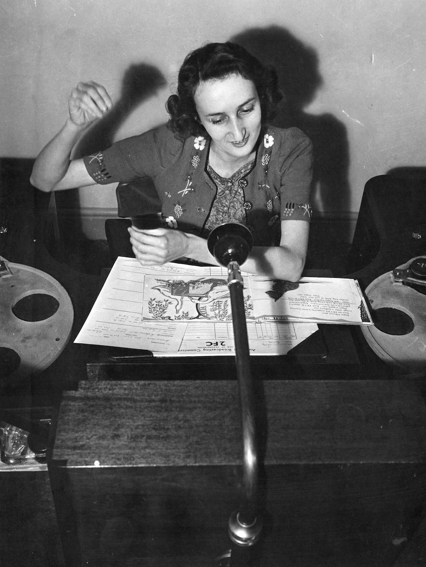 A woman sits at a desk and reads from papers into a microphone.