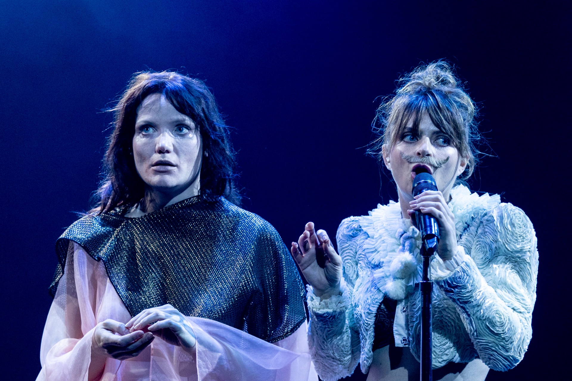 Two female actors performing on stage; one on the right wears a fake moustache and is holding a microphone to her mouth