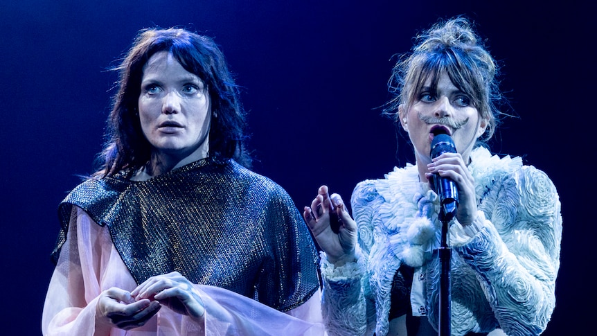 Two female actors performing on stage; one on the right wears a fake moustache and is holding a microphone to her mouth