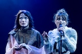 Two female actors performing on stage; one on the right wears a fake moustache and is holding a microphone to her mouth