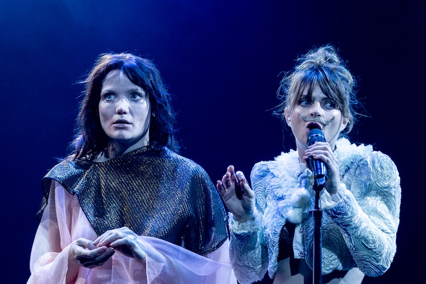 Two female actors performing on stage; one on the right wears a fake moustache and is holding a microphone to her mouth