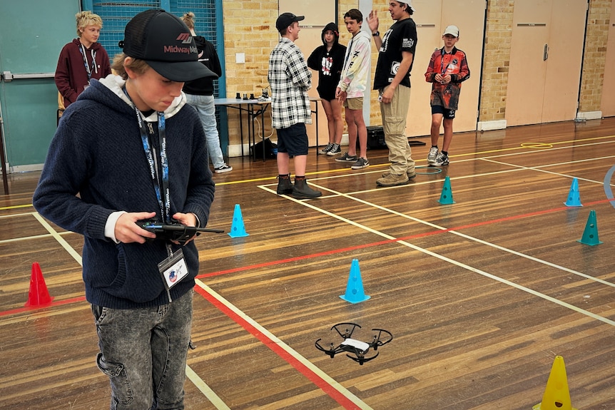 Blond boy wearing black cap, lanyard, black top, operating drone in a school gym, other children stand behind talking.