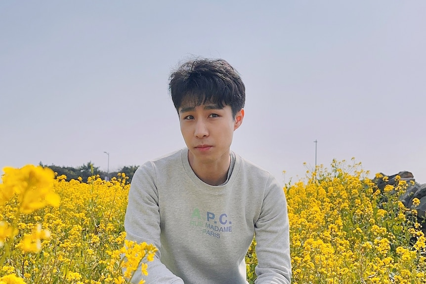 Michael Ko is squatting down in a field of yellow flowers, he's wearing a grey sweatshirt and blue jeans.