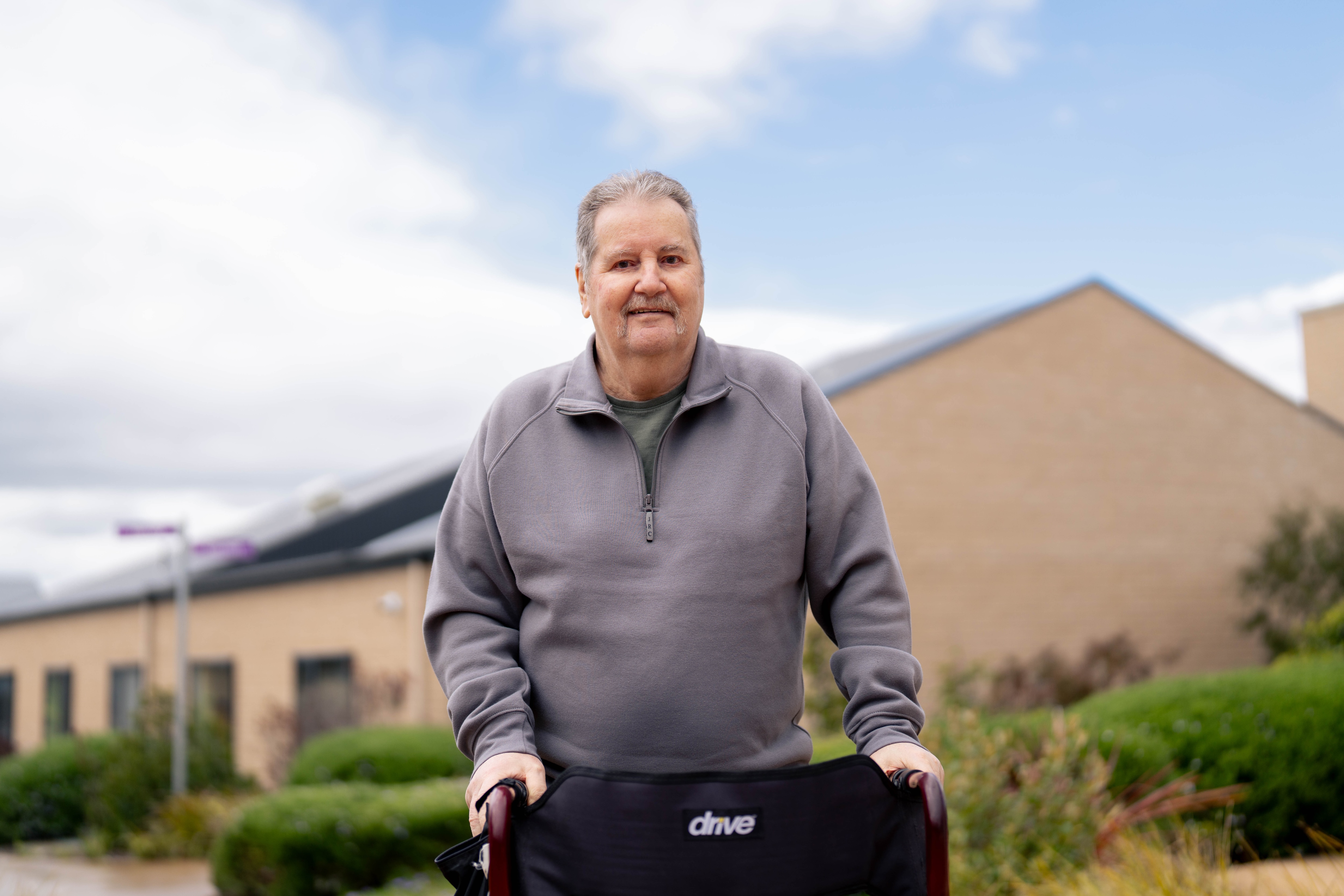 Nick, wearing a grey quarter zip jacket, stands with a walker smiling for a photo with aged care housing behind him.
