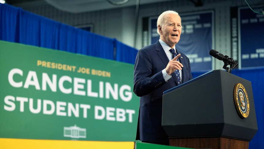 Joe Biden stands at a lectern, pointing, in front of a sign saying CANCELING STUDENT DEBT