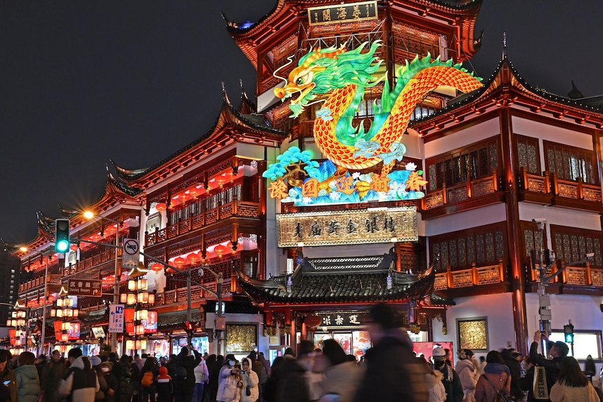 A crowd of people outside a temple with a light up dragon. 
