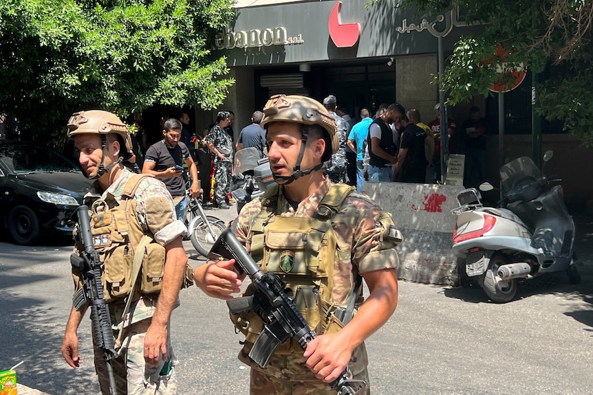 Dos soldados con cascos y uniformes de camuflaje y rifles están parados en una calle frente a una multitud de hombres.