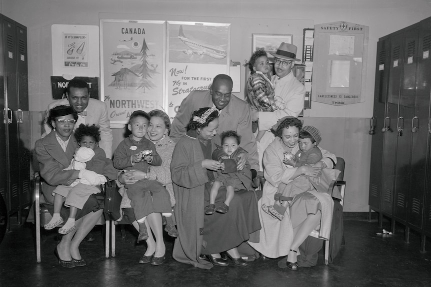 Black and white 1956 photo four couples with five children. Travel posters behind.