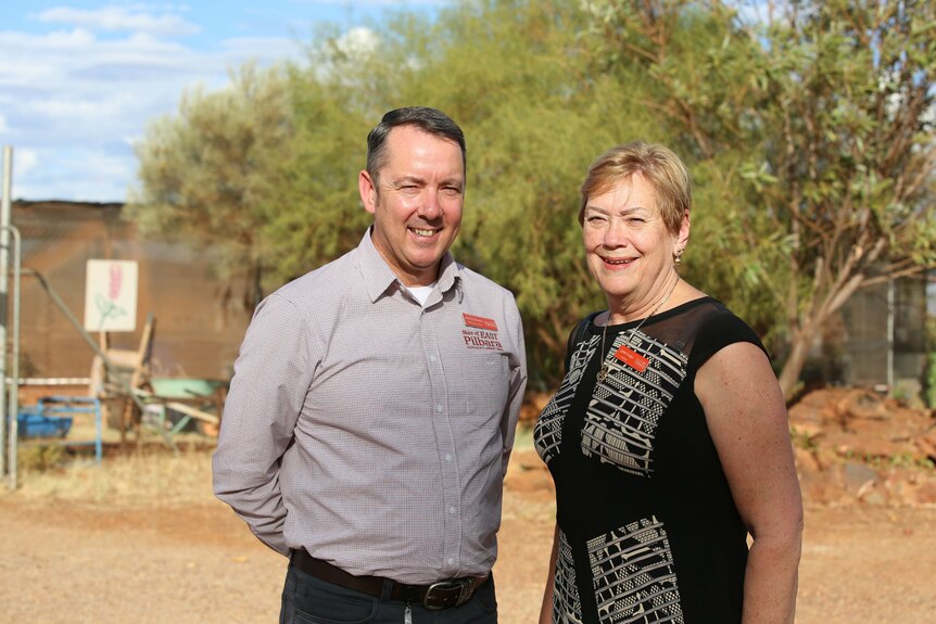 A man and a woman stand together smiling, squinting in the sun, in front of trees and red dirt.