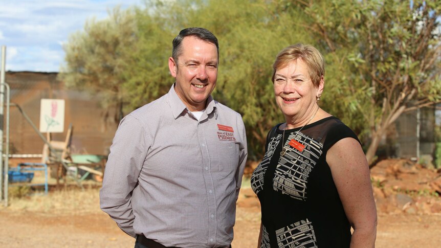 A man and a woman stand together smiling, squinting in the sun, in front of trees and red dirt.