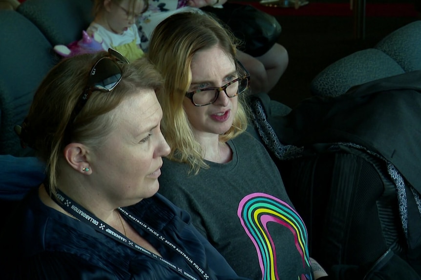 A woman wearing glasses speaking to another woman wearing a lanyard at an airport chair
